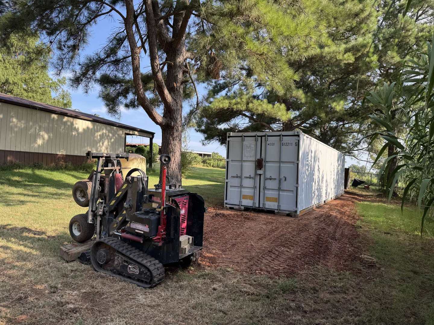 White container placed beside a pine tree with a crawler mover
